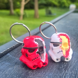 Puede incluir: Dos llaveros con cascos de Stormtrooper de Star Wars con un diseño de calavera. Un casco es rojo y negro, el otro es blanco con una calavera amarilla.