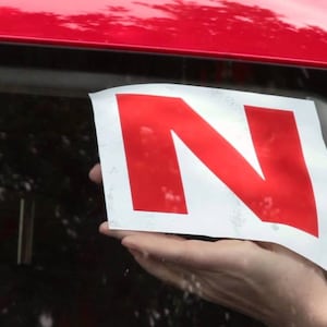 Op de afbeelding: Een wit bord met een rode letter N erop. Het bord wordt tegen de achterruit van een rode auto gehouden.