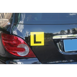 May include: A black car with a yellow L-shaped sign on the back. The sign indicates a learner driver.