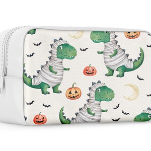 May include: White zippered pouch with a Halloween-themed print featuring green mummies dressed as dinosaurs, orange pumpkins, and black bats.