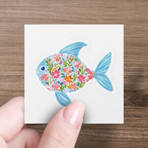 Sticker of a fish filled with colorful watercolor flowers, held by a hand.