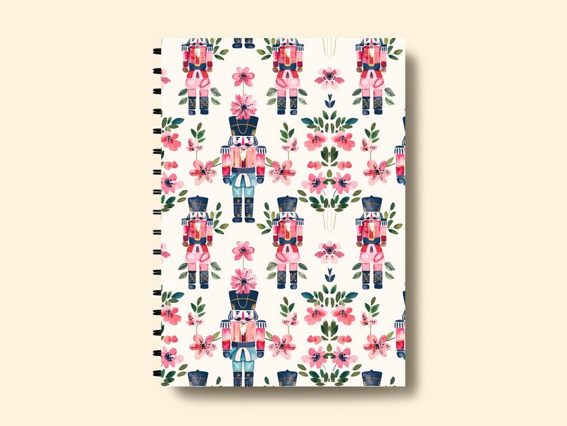 May include: A white spiral bound notebook with a repeating pattern of pink flowers and watercolor nutcracker soldiers.