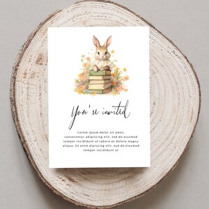 Op de afbeelding: Een witte kaart met een aquarel-illustratie van een konijn dat op een stapel boeken zit, omringd door bloemen. De tekst "You're invited" is geschreven in zwarte cursieve letters. De kaart heeft een witte achtergrond en de tekst "Lorem ipsum dolor sit amet, consectetur adipiscing elit, sed do eiusmod tempor incididunt ut labore et dolore magna aliqua. Ut enim dipiscing elit, sed do eiusmod tempor incididunt ut" is in zwart geschreven.