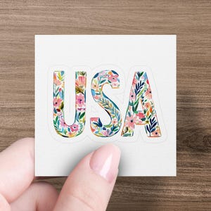 Sticker with floral letters spelling "USA" held by a hand.