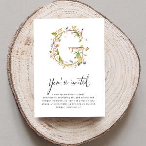 May include: A white invitation card with a floral wreath in shades of green, yellow, and pink forming the letter "G". The text "You're invited" is written in black cursive script below the wreath. The text "Lorem ipsum dolor sit amet, consectetur adipiscing elit, sed do eiusmod tempor incididunt ut labore et dolore magna aliqua. Ut enim dipiscing elit, sed do eiusmod tempor incididunt ut" is written in black below the cursive text.