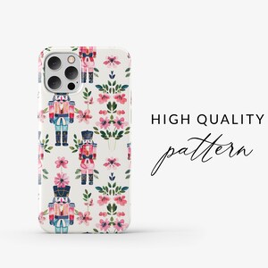 May include: A white phone case with a repeating pattern of pink flowers and nutcracker soldiers. The case has a cutout for the phone's camera.