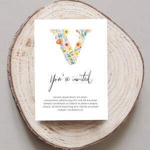 May include: A white card with a floral letter V and the text "You're invited". The card is on a wooden surface.
