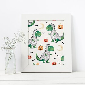 May include: A white framed print with a repeating pattern of green mummies, orange pumpkins, and black bats on a white background. The mummies are dinosaurs wearing white bandages.