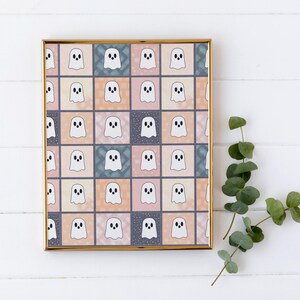 May include: A framed print with a repeating pattern of cartoon ghosts in a grid. The ghosts are white with black eyes and mouths, set against pastel squares in peach, blue, and grey. A sprig of green eucalyptus is in the corner.