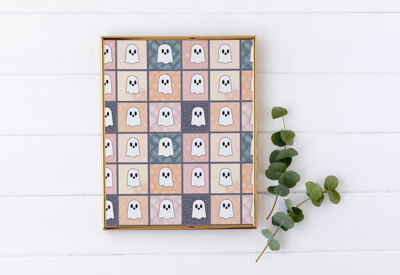 May include: A framed print with a repeating pattern of cartoon ghosts in a grid. The ghosts are white with black eyes and mouths, set against pastel squares in peach, blue, and grey. A sprig of green eucalyptus is in the corner.