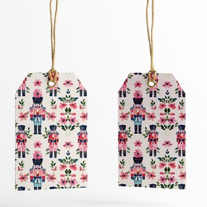 May include: Two white gift tags with a repeating pattern of pink flowers and nutcracker soldiers. The tags are tied with twine.