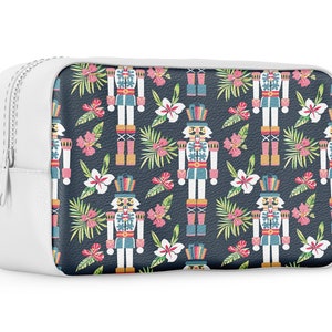 Pu&ograve; includere: Borsa bianca con cerniera con sfondo blu e un motivo ripetuto di figure di schiaccianoci, foglie tropicali e fiori rosa.