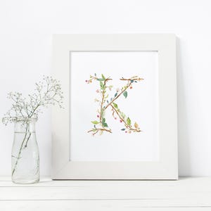May include: A white frame with a white mat surrounding a print of the letter K made of green leaves and pink flowers.