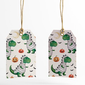 May include: Two white gift tags with a green dinosaur mummy pattern. The pattern also includes orange pumpkins, bats, and a crescent moon. The tags are tied with twine.