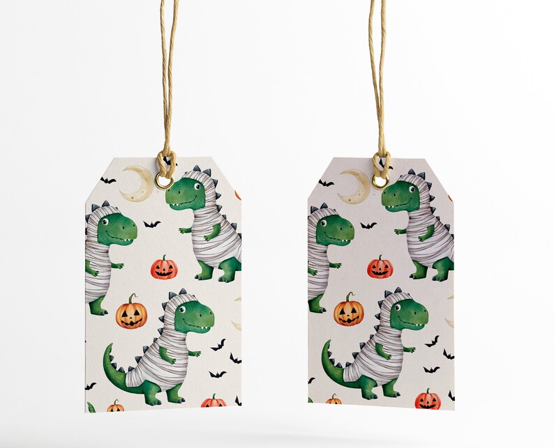 May include: Two white gift tags with a green dinosaur mummy pattern. The pattern also includes orange pumpkins, bats, and a crescent moon. The tags are tied with twine.