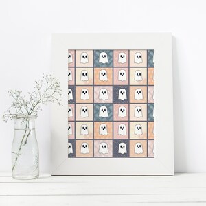 May include: A framed print with a repeating pattern of white cartoon ghosts on a patchwork of pastel squares. The squares are in shades of pink, peach, blue, and grey. The print is in a white frame, and a glass vase with white flowers is in the foreground.