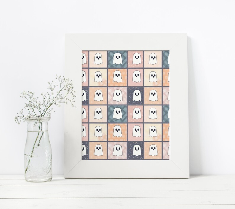 May include: A framed print with a repeating pattern of white cartoon ghosts on a patchwork of pastel squares. The squares are in shades of pink, peach, blue, and grey. The print is in a white frame, and a glass vase with white flowers is in the foreground.