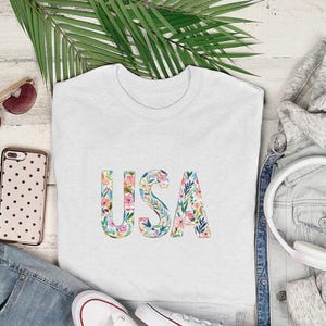 White t-shirt with floral "USA" graphic, styled with jeans and headphones.