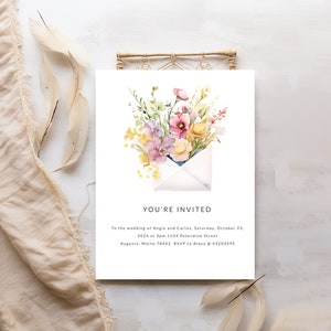 May include: A wedding invitation featuring a watercolour illustration of a white envelope with a bouquet of pink, yellow, and purple flowers inside. The text reads "You're Invited" and includes the wedding details: Angie and Carlos, Saturday, 23rd October, 2024 at 3pm, 1234 Peterwise Street, Augusta, Maine 78402. RSVP to Alexa @ 43203495.