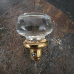 May include: A clear glass knob with a gold metal base. The knob is attached to a wall with a screw.