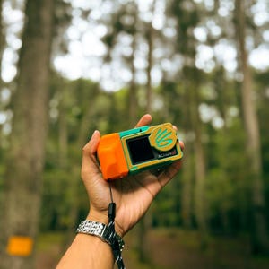 May include: An orange and teal action camera with a black screen and the word "ACTION" printed on the side. The camera is held in a hand with a silver watch and a black and white lanyard. The background is a blurred forest.