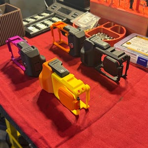 May include: Several 3D-printed devices in various colors, including yellow, orange, and purple, are arranged on a red surface. Each device has a black component and a unique frame. Other tools and components are visible in the background.