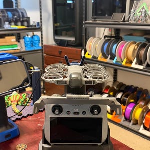May include: A grey drone with four propellers sits above a grey remote control with a black screen, on a black base. The background shows shelves with filament spools and 3D printers.
