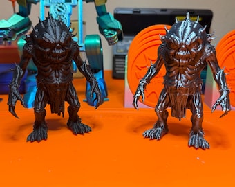 May include: Two detailed, 3D-printed monster figurines. One is black, the other is silver. Both have spiky heads, clawed hands, and feet. They stand on an orange surface, with a partially visible robot and other objects in the background.