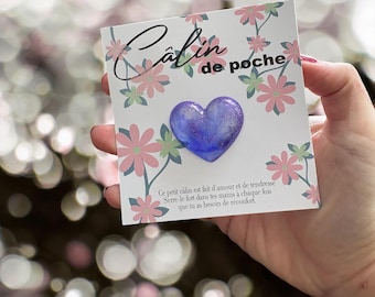 May include: A small, heart-shaped, blue and purple pocket hug. The heart is attached to a card with floral designs and the words "Calin de poche". The card also has a message in French.