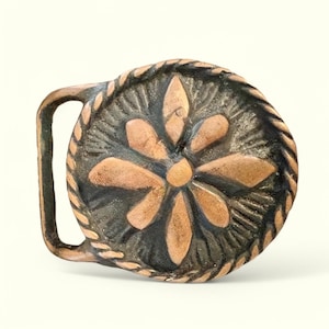 May include: A copper-colored belt buckle with a detailed design of a flower with a center circle. The buckle is surrounded by a rope-like border.