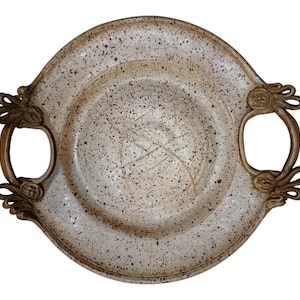 Puede incluir: Una fuente de cerámica moteada de color blanquecino con dos asas de bronce ornamentadas. La fuente tiene un borde ligeramente elevado y una superficie texturizada con motas marrones oscuras. Las asas presentan diseños florales decorativos.