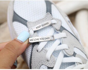 May include: Close-up of a white and gray athletic shoe with white laces. Silver metal shoelace tags read "KEEP GOING!" and "WE LOVE YOU DAD!" The shoe is being held by a hand with a light blue painted fingernail.