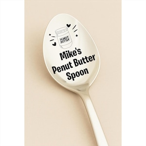 May include: A stainless steel spoon with the words "Mike's Peanut Butter Spoon" in black text. Above the text is a graphic of a peanut butter jar with the words "Peanut Butter". The spoon has a shiny, reflective surface.