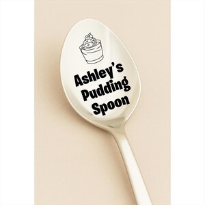 May include: A silver spoon with the text "Ashley's Pudding Spoon" in black. Above the text is a small illustration of a dessert in a cup. The spoon is on a light beige background.