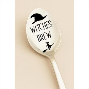 May include: A silver spoon with the words "WITCHES BREW" in black lettering. Above the text is a black witch hat, and below is a silhouette of a witch on a broom. The spoon is on a light beige background.