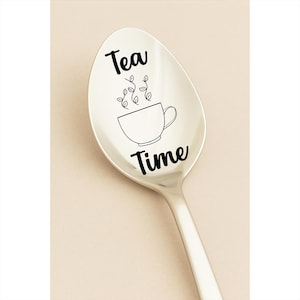 May include: A silver spoon with the words "Tea Time" in black script. A line drawing of a teacup with steam rising from it is centered on the spoon's bowl. The spoon is set against a light beige background.