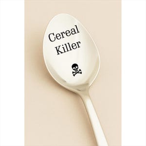 May include: A stainless steel spoon with the words "Cereal Killer" in black, along with a skull and crossbones graphic. The spoon has a polished finish and a long handle.