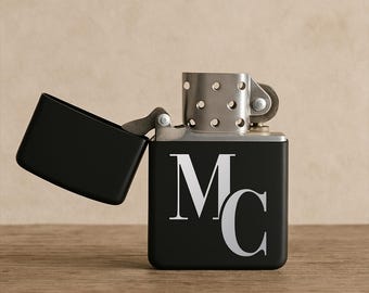 May include: A black lighter with the lid open, revealing a silver interior. The lighter features the silver initials "MC" on the front. The lighter is set against a wooden surface and a neutral background.
