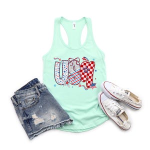 May include: A light green tank top with the word "USA" in a patriotic design, paired with denim shorts and white sneakers. The "USA" graphic features stars, stripes, and a checkered pattern in red, white, and blue.
