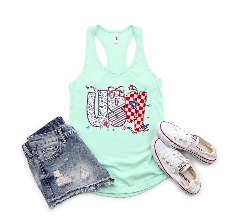 May include: A light green tank top with the word "USA" in a patriotic design, paired with denim shorts and white sneakers. The "USA" graphic features stars, stripes, and a checkered pattern in red, white, and blue.