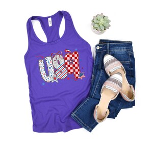 May include: A purple racerback tank top with the word "USA" in a patriotic design, paired with blue jeans and striped flats. The "USA" graphic features stars, stripes, and a checkered pattern.