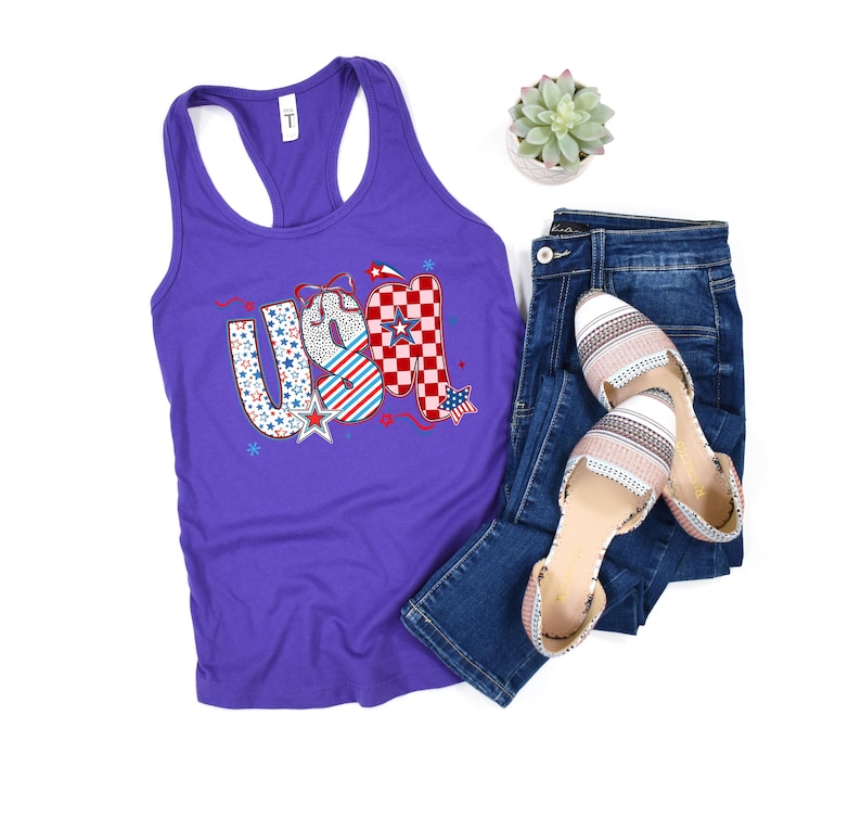 May include: A purple racerback tank top with the word "USA" in a patriotic design, paired with blue jeans and striped flats. The "USA" graphic features stars, stripes, and a checkered pattern.