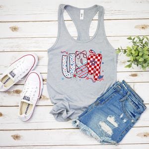 May include: A gray tank top with the word "USA" in red, white, and blue patterns, paired with denim shorts and white sneakers. The patriotic design includes stars and stripes, perfect for celebrating the Fourth of July.