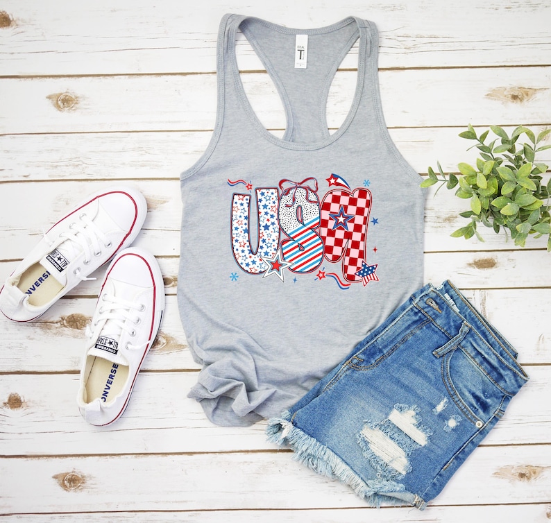 May include: A gray tank top with the word "USA" in red, white, and blue patterns, paired with denim shorts and white sneakers. The patriotic design includes stars and stripes, perfect for celebrating the Fourth of July.