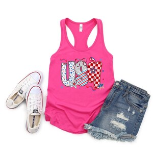 May include: A hot pink tank top with the word "USA" in a patriotic design, paired with denim shorts and white Converse sneakers. The "U" is blue with white stars, the "S" is white with red and blue stripes, and the "A" is red and white checkered.