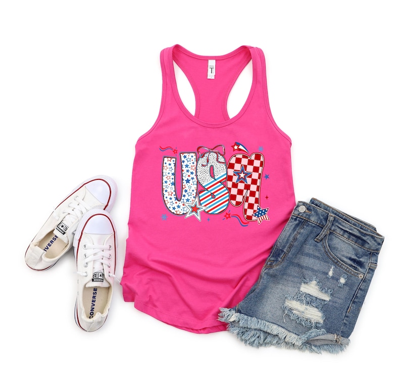May include: A hot pink tank top with the word "USA" in a patriotic design, paired with denim shorts and white Converse sneakers. The "U" is blue with white stars, the "S" is white with red and blue stripes, and the "A" is red and white checkered.