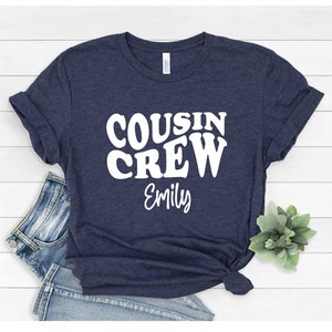 May include: A navy blue t-shirt with the text "COUSIN CREW" in white, retro-style lettering. The name "Emily" is written below in white.