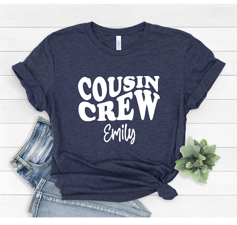 May include: A navy blue t-shirt with the text "COUSIN CREW" in white, retro-style lettering. The name "Emily" is written below in white.