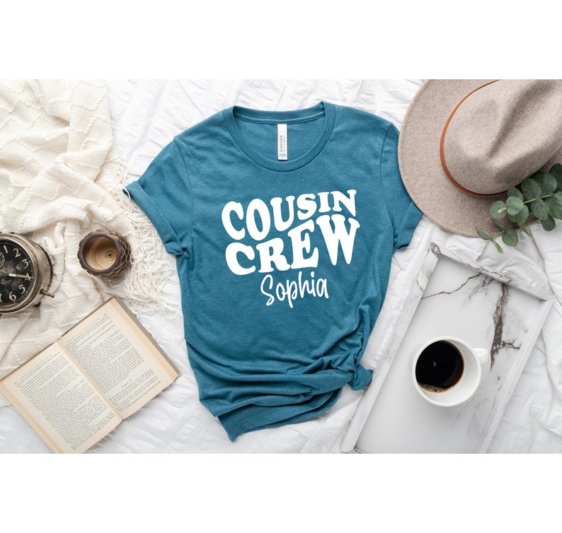 May include: A teal blue t-shirt with the text "COUSIN CREW" in white retro-style lettering. The name "Sophia" is written below in white.