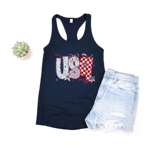 May include: A navy blue tank top with the word "USA" in a patriotic design. The letters feature patterns of stars, stripes, and checkered elements in red, white, and blue. Light blue denim shorts are next to the tank top.
